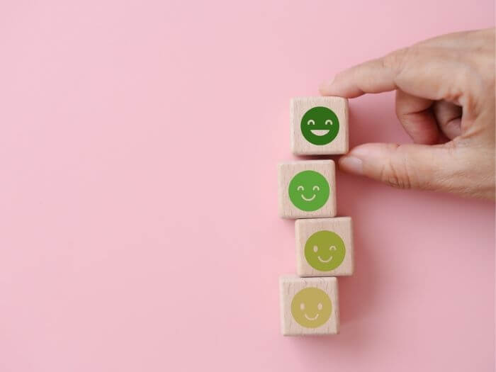 Four stacked building blocks against a pastel pink background, each has a green smiley face which gets brighter and more happy the higher it is stacked.