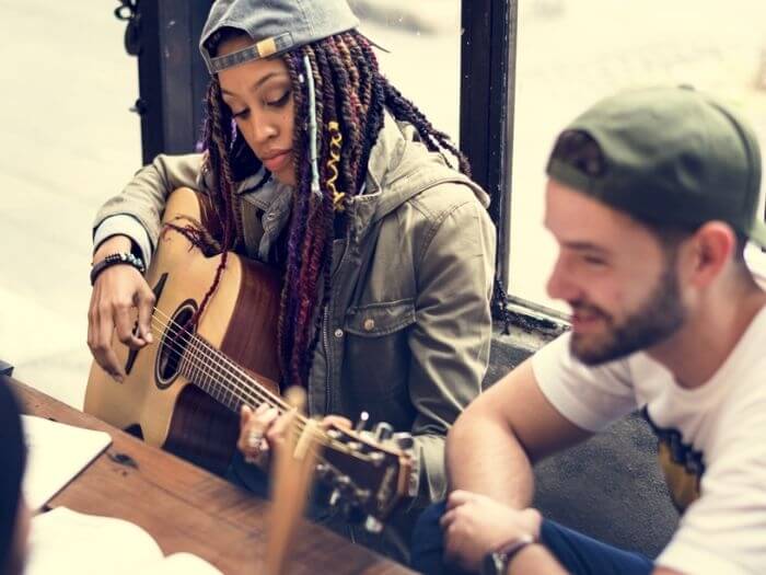 Woman playing acoustic guitar at a table with male musician, paper and pen are on the table next to them.