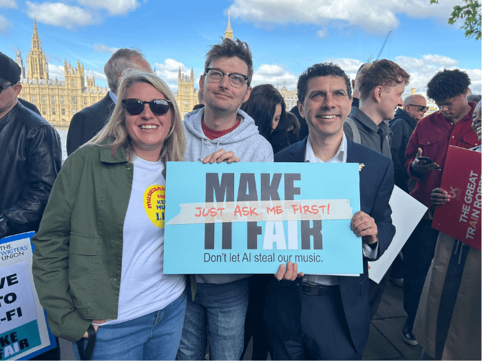 Naomi Pohl holding a sign saying 'Don't let AI steal our music - make it fair' in front of Parliament with a large demo group.