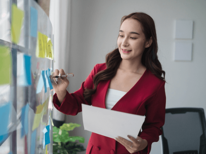 A woman in a red jacket writing on post-it notes on a board at work.