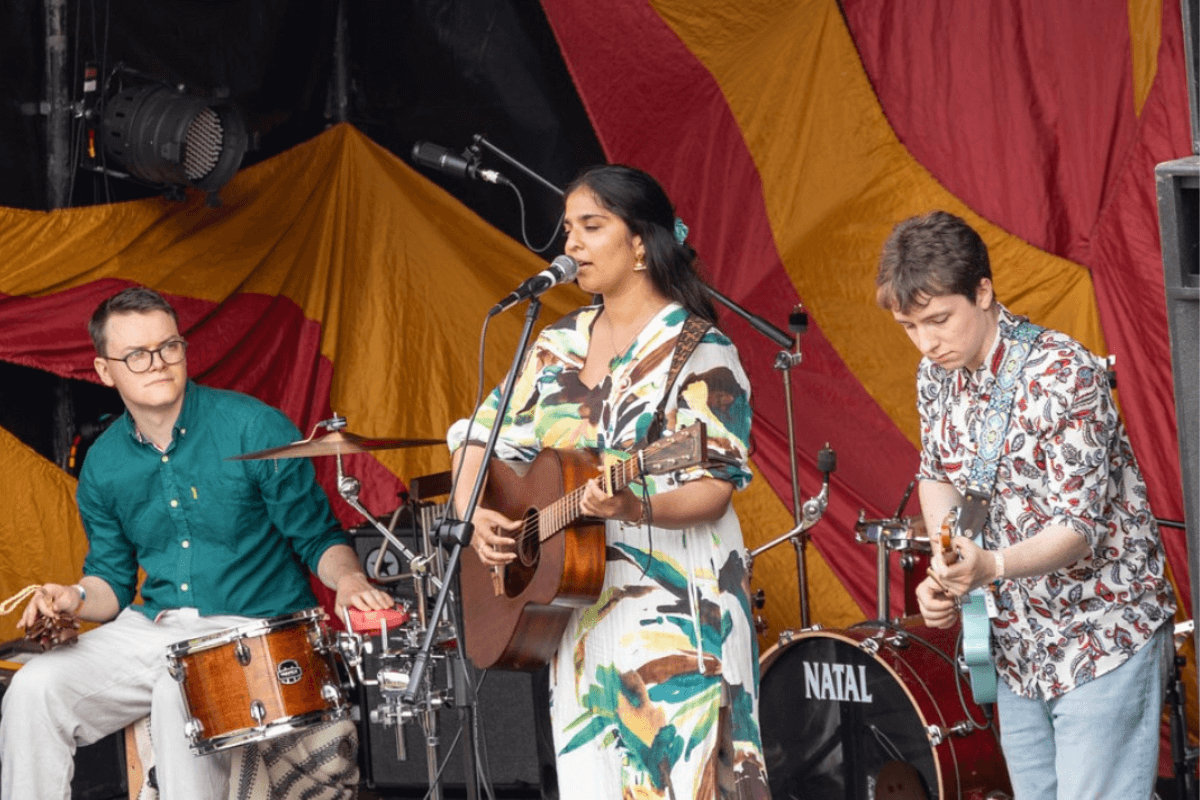 Aayushi on stage playing guitar and singing into a microphone, a drummer is to her left, and another guitarist to her right.