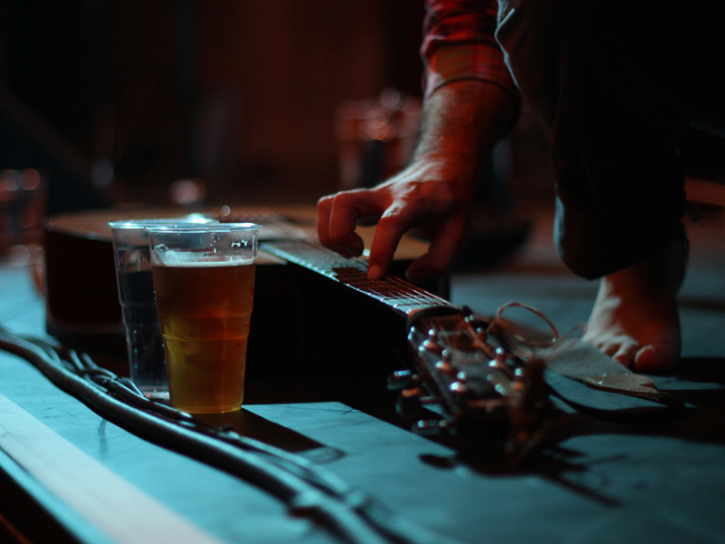 Photograph of a small stage set up, we can see an acoustic guitar placed on the stage floor, alongside a half empty pint of beer. A mans hand reaches down to touch the strings.