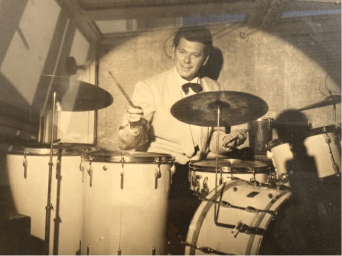 A sepia photograph of a young Alfred playing drums, dressed in a white suit with bow tie, smilig to camera.