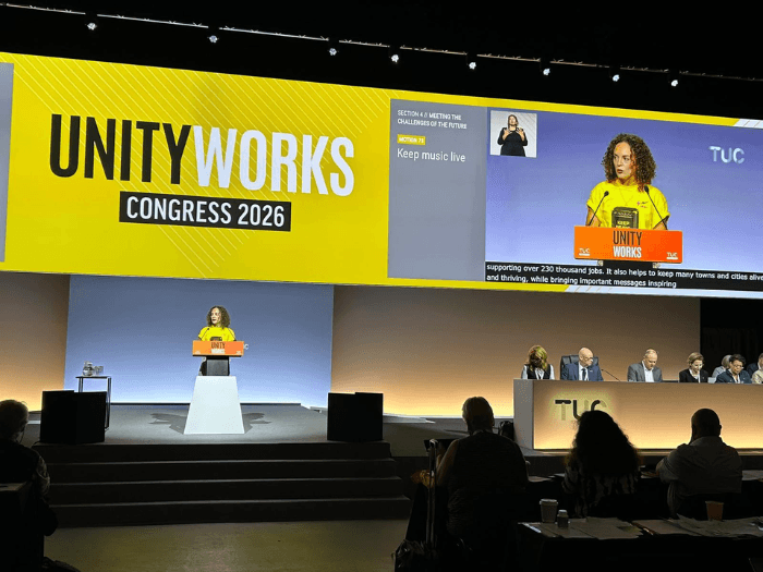 Alice Angliss on stage in front of a lectern, behind her is a yellow coloured screen showing a video of her with a person using sign lanage in the corner. Text says: "Unity Works Congress 2026".