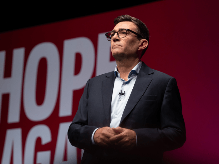 Andy Burnham wearing a suit standing against a red backdrop with white lettering.