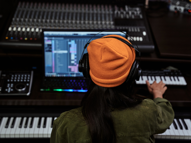 Woman in a recording studio facing away from the camera, one hand on the keyboard while looking at the laptop.