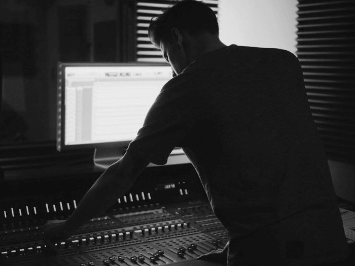 Black and white image of young man over a mixing desk with a large PC monitor screen in front of him.