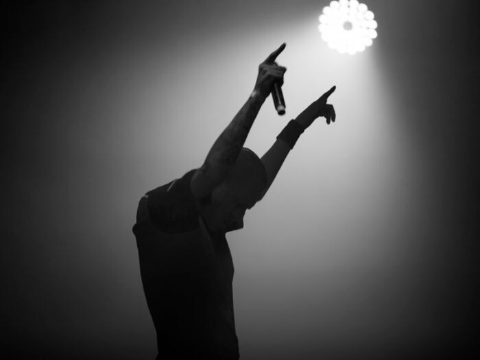 Black and white silhouette of singer under a spotlight with both arms raised and a  microphone in one hand.