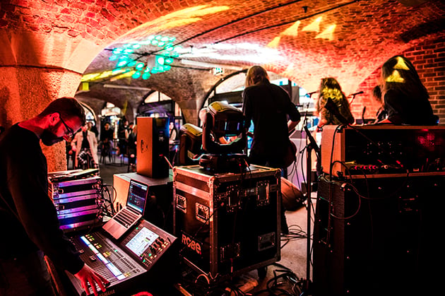 Photograph of a band playing, in what appears to be underground archways, with red lights shining.