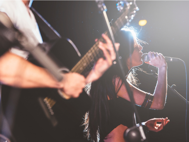 Musicians playing in a band setting, we can see a woman singing into a microphone and a man playing on the guitar