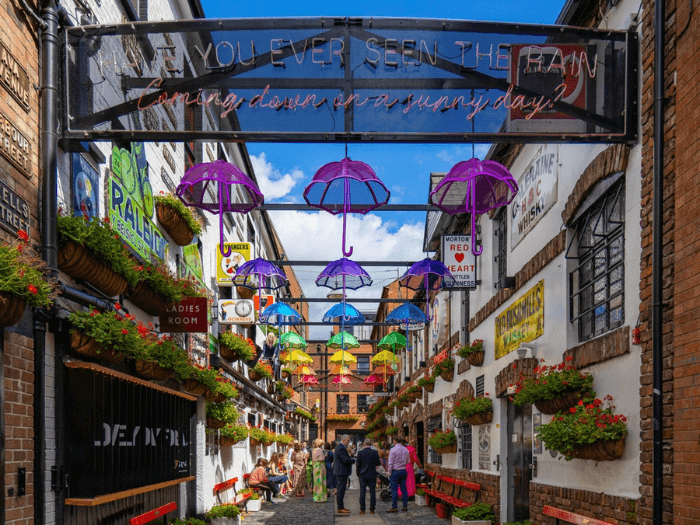 A famous, vibrant street in Commercial Court in the Cathedral Quarter of Belfast. The street is lined with historic pubs, benches and flowers, with umbrellas and neon signs hanging over the alleyway.