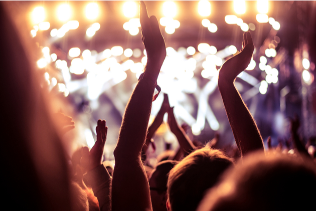 Close up of a group of people in soft focus and dim lighting at a concert, their hands are in the air as if they are mid clap and the stage is lit with out of focus lights.