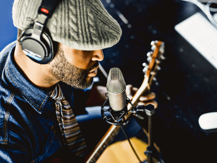 Black male musician wearing cap and headphones sat in recording studio with mic and acoustic guitar.