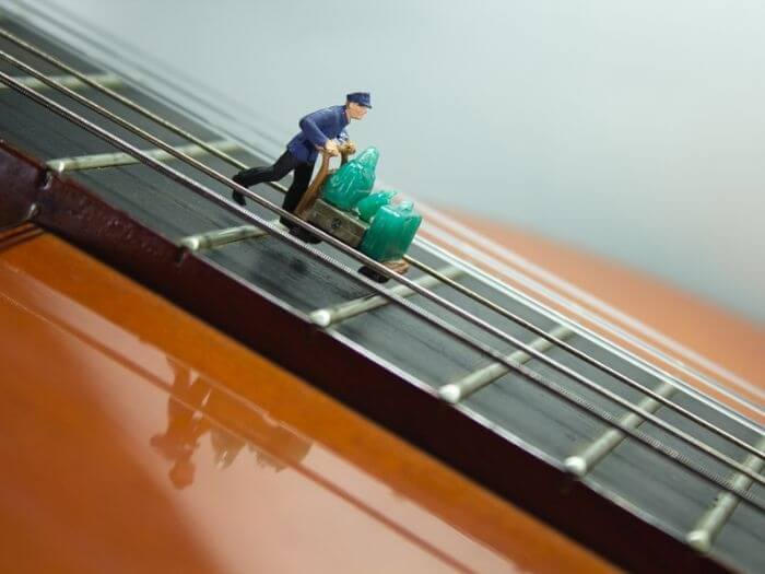 A miniature figurine of a porter with a full luggage cart, placed on the neck of an acoustic guitar. Instrument and baggage concept.