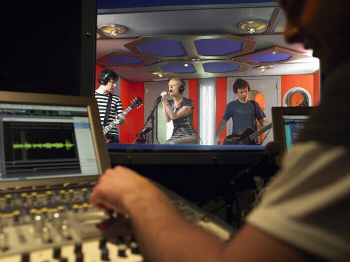 Band in recording studio booth, with technician in foreground by mixing desk.