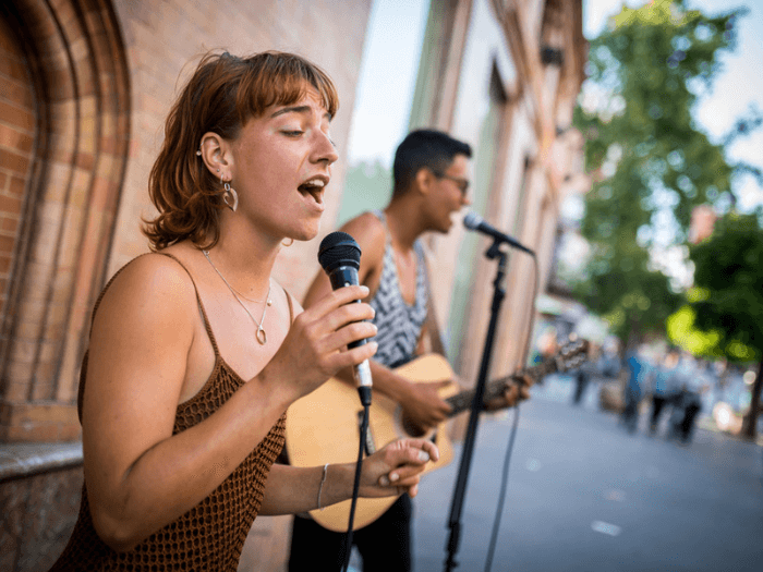 Two young buskers, one singing and one playing guitar on a sunny street.