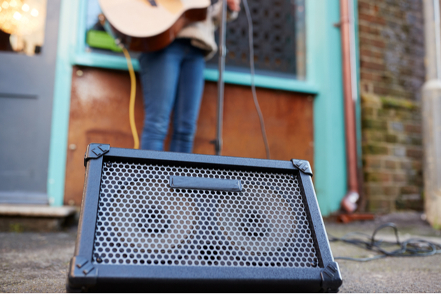 Photograph of a busker playing outdoors on a guitar on a busy street, the focus is on the amplifier with the performer blurred in the background.
