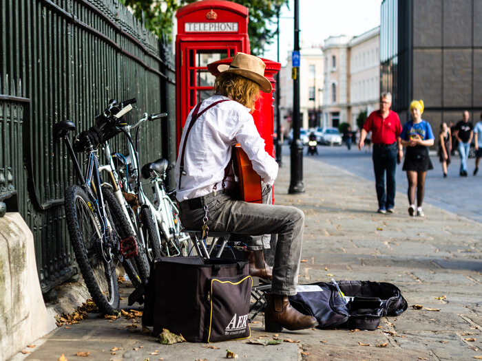 Busker sat on a small camping stall in London next to a red phone box, playing acoustic guitar on his knee with a guitar case open for passers-by.