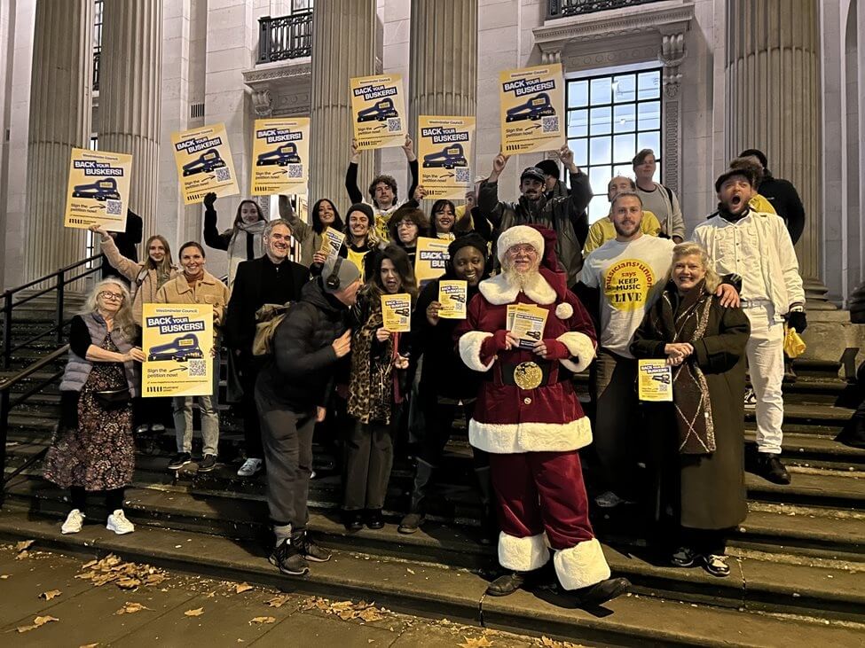A group of buskers standing on the steps of Westminster Council holding campaign placards.