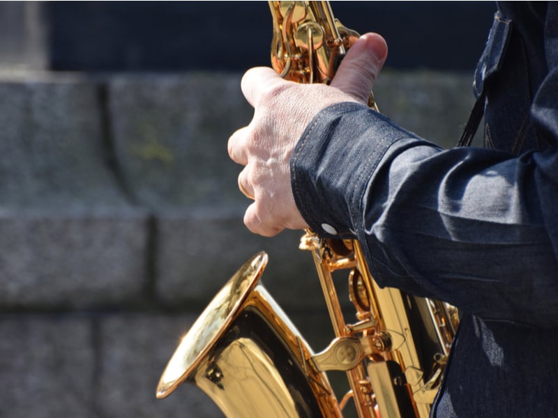 Photograph of a saxophone up close, the background of a brick outdoor wall implies that the performer is outside, we can only see the performers arm and hand.