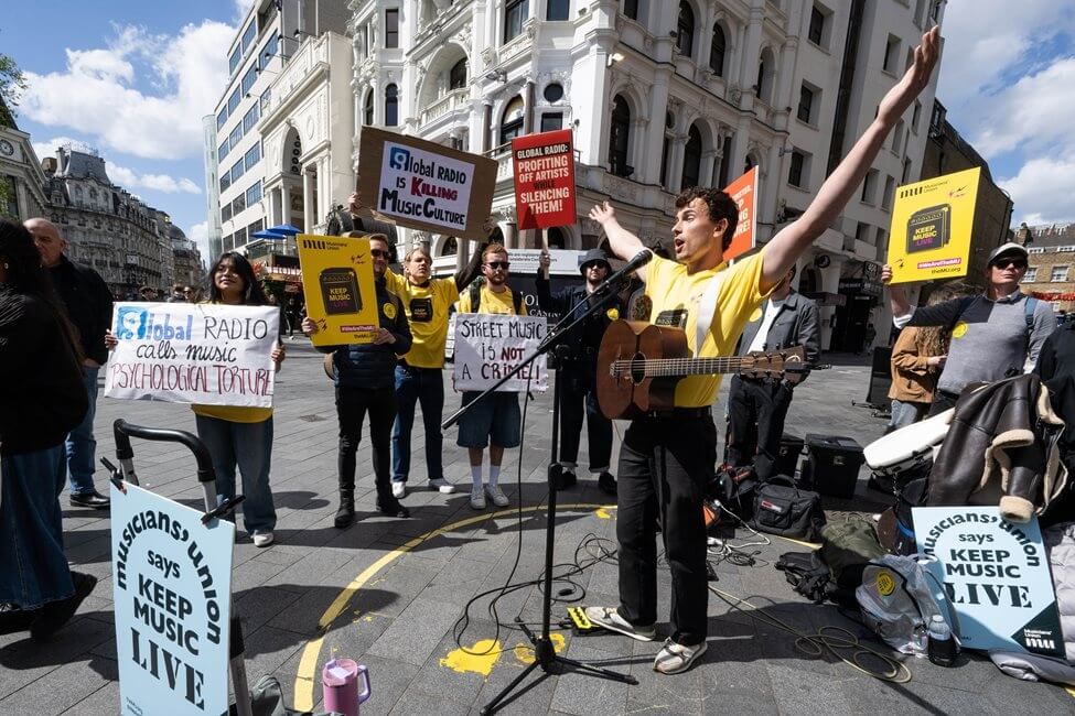 A busker wearing a yellow Musicians' Union t-shirt surrounded by supporters with signs saying 'keep music live' at a busking demo in London in April.