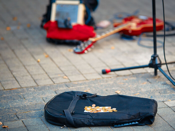 A guitar case lies in the street with a pool of coins on it, in the background is a mic stand, a guitar and an amp.