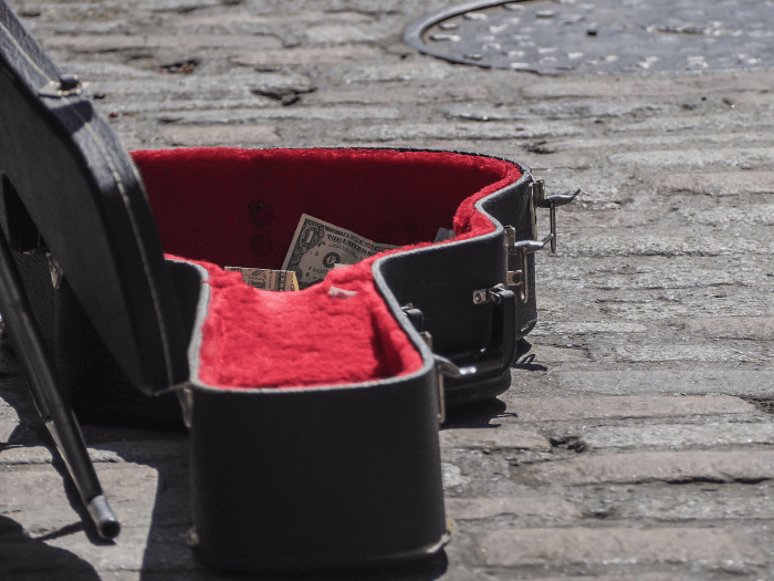 Dollar bills visible in busker's guitar case on cobbled street.