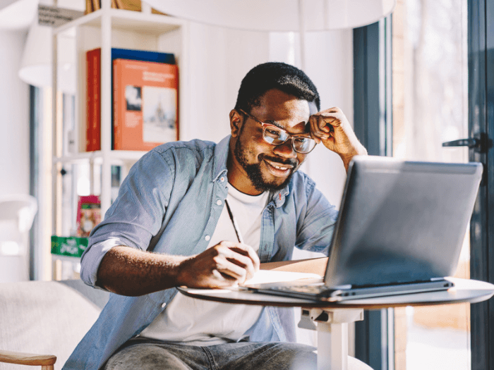 Young man, wearing glasses, at home smiling while taking notes and looking at a laptop.