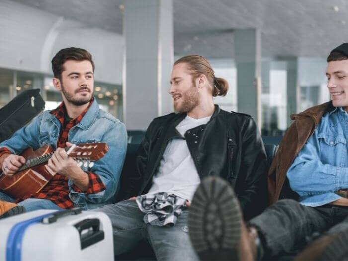 Three people sat in an airport with suitcase and an acoustic guitar.