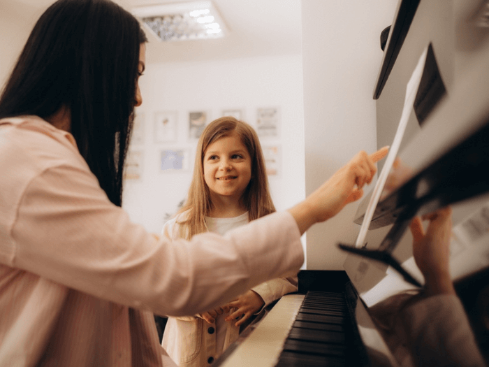 A female music teacher sitting at a piano with a young girl teaching her how to play.