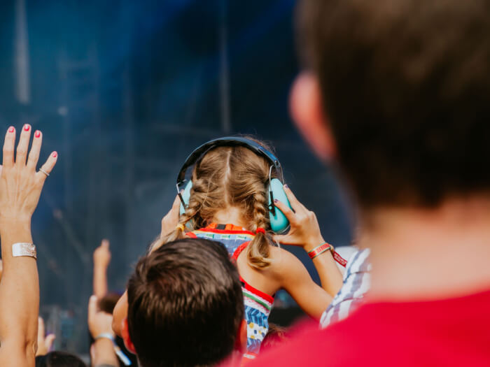 Small child wearing ear defenders sits on someone's shoulders while looking at a large concert at a festival