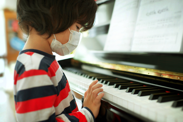 Photograph of a small child sat wearing a facemask in front of a piano keyboard.