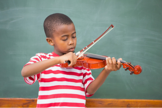Photograph of a young child standing in front of a chalk board, learning the violin.