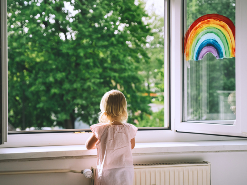 Photograph of a small child looking out a window onto a Spring like scene, a rainbow has been painted on the glass.