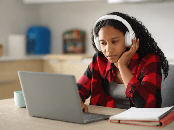 Young Black college-age woman, sat at a kitchen  table with headphones on, looking at a laptop screen in concentration, next to a stack of books and notes.