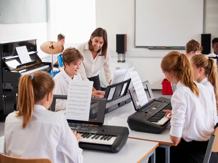 Secondary school children sat around tables in a classroom, sharing an electronic keyboard having a music lesson.