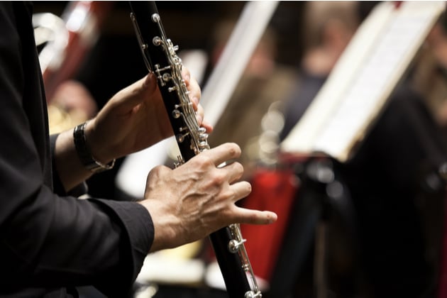 Photograph of a clarinet performer in what appears to be an orchestral setting, although we can only see the performers hands and a blur of other instruments and music stands in the background.