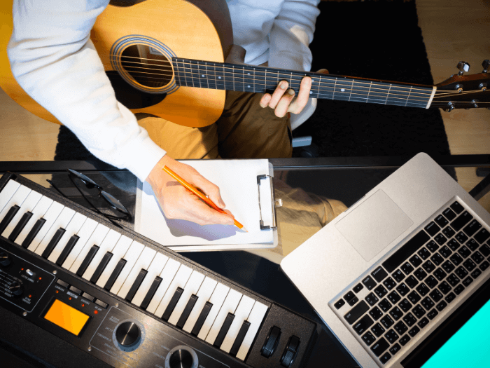 Birds eye view of person in a blue shirt holding a pencil making notes while a guitar is balanced on their knee, with an open laptop and keyboard next to them, representing composing a song.
