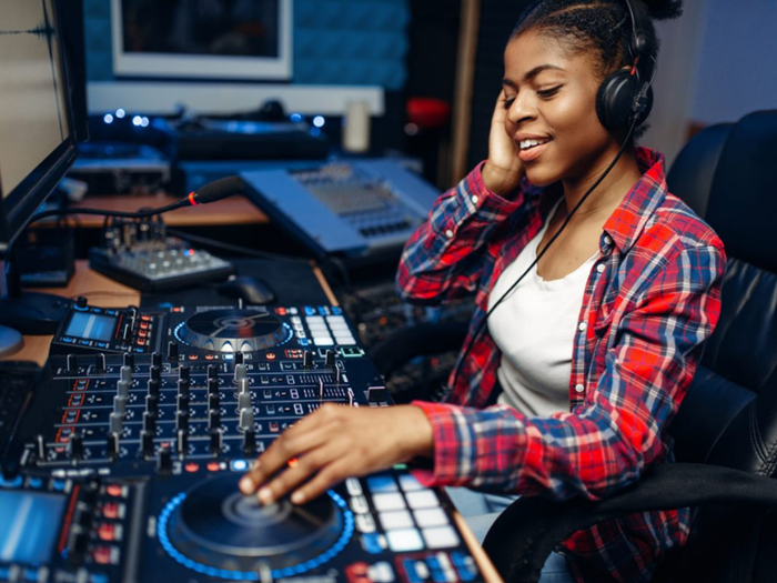Girl in red checked shirt wearing over ear headphones, one hand to one ear, also looking down at a studio mixing desk