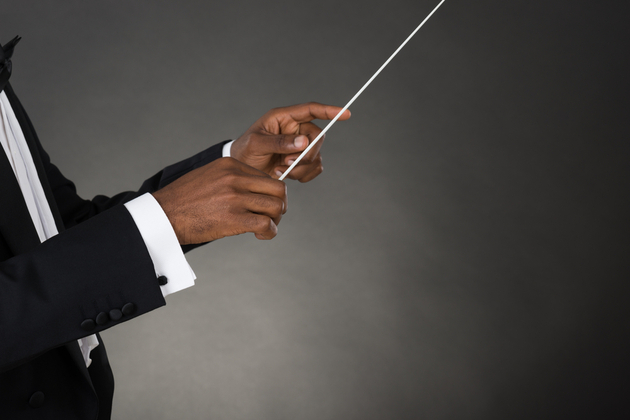 Photograph of a Black man conducting - holding a raised conductors batton, with the photograph focusing on his hands.