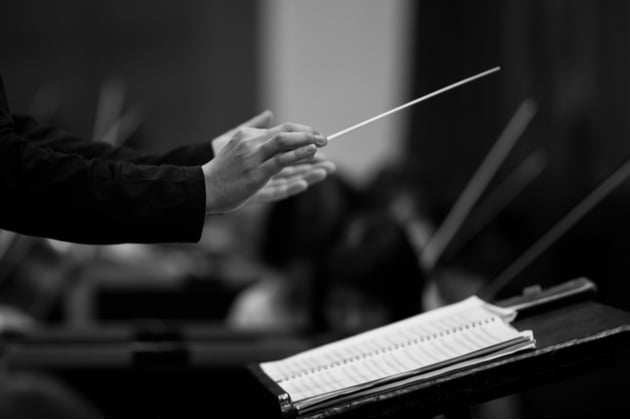 Black and white photograph of a conductors hands, holding a baton over the music stand at the head of an orchestra. The musicians in the background are blurred.