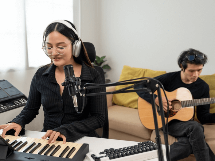 Two young musicians in a home studio, one at a desk recording on keyboard with microphone, the other sat behind her playing an acoustic guitar.