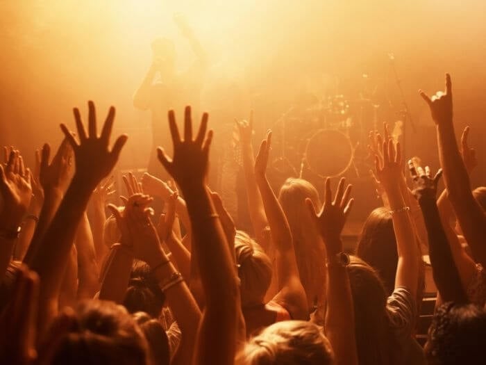 Audience right in front of a stage at a gig, hands raised in the air. Band are on stage, cast in an orange glow from the stage lights.