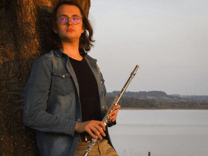 Daniel stood against a tree at sunset, holding a flute, with a forest and lake in the background.