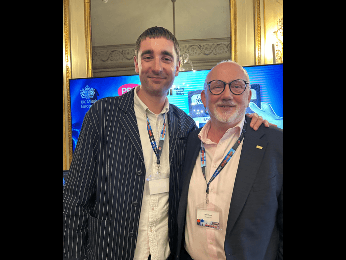 Two men standing for a photograph together at a work event. They are smiling and wearing matching lanyards.