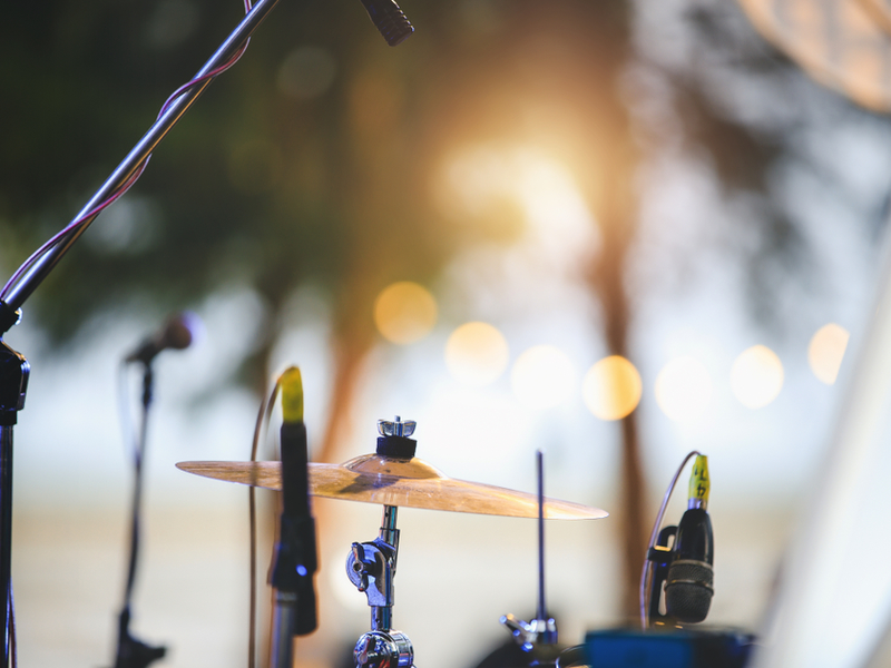 Photograph of a drum kit against a blurred background, which appears to be the outdoors. A string of brightly coloured lights suggests that this could be a festival.