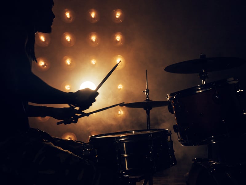 Photograph of a drummer playing in silhouette against smoke and bright golden light.
