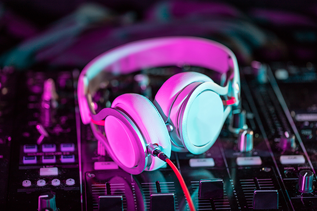 Photograph of a pair of white earphones resting on a mixing desk. There is a strange pinky-blue light shining over the earphones from the left-hand side.