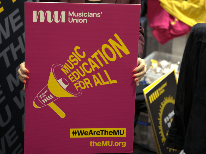 Close up of a pink board being held up, which says “Music Education For All’ coming from a megaphone drawing in yellow, with the Musicians’ Union logo.