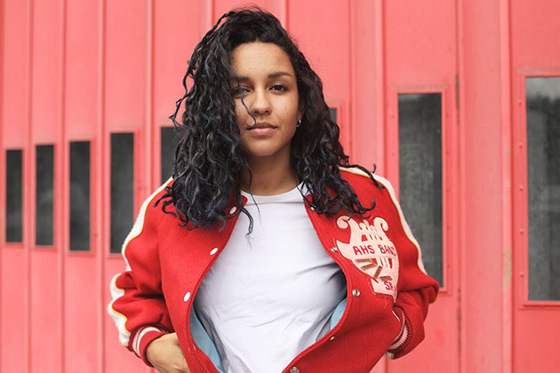 Photograph of Scottish-Sudanese singer/songwriter Eliza Shadad wearing a red jacket and standing in front of a red background.